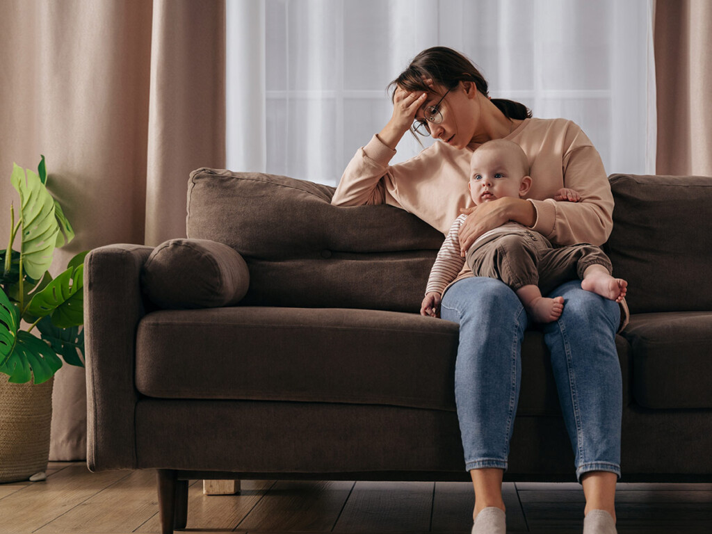 Image to depict gender inequality: a stressed out looking mother sits on a couch holding a baby