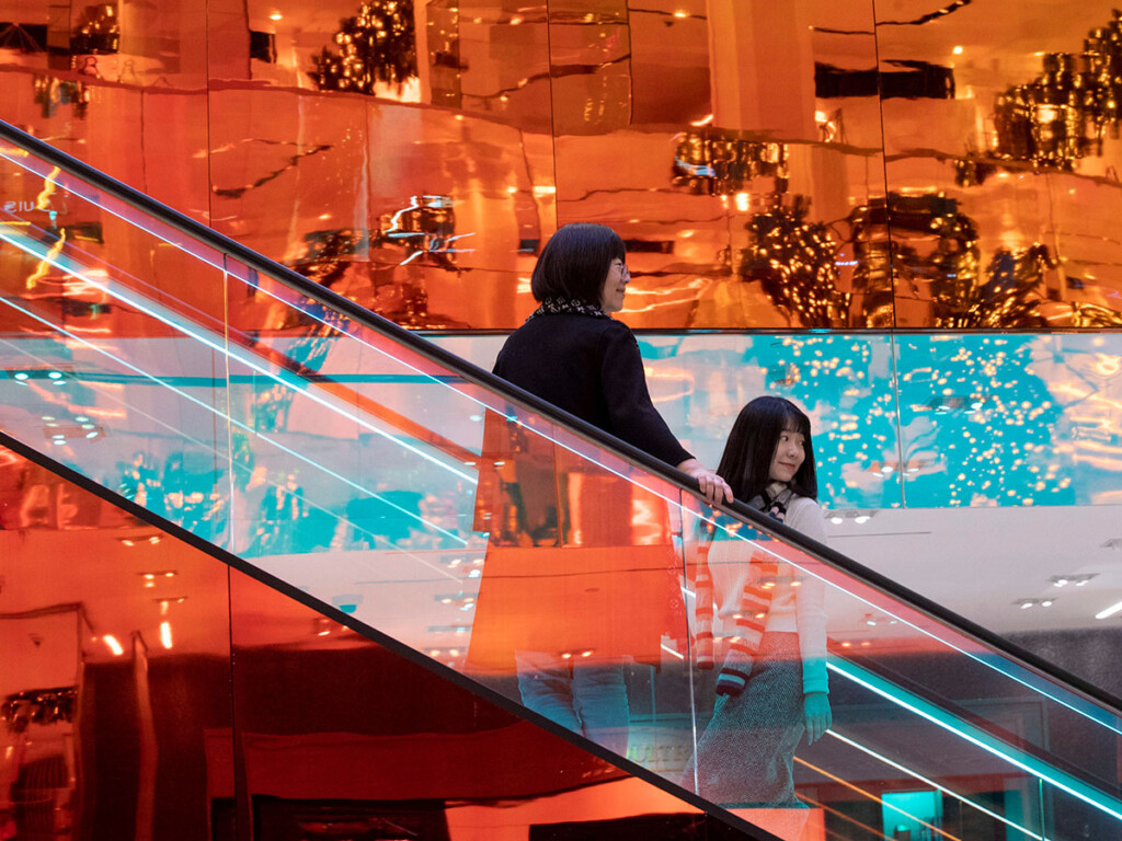 Two holiday shoppers descend on an escalator in what looks to be a mall.