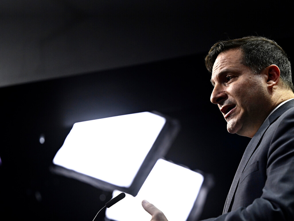 A sideview of a man speaking while at a podium with a bright light shining in the background.