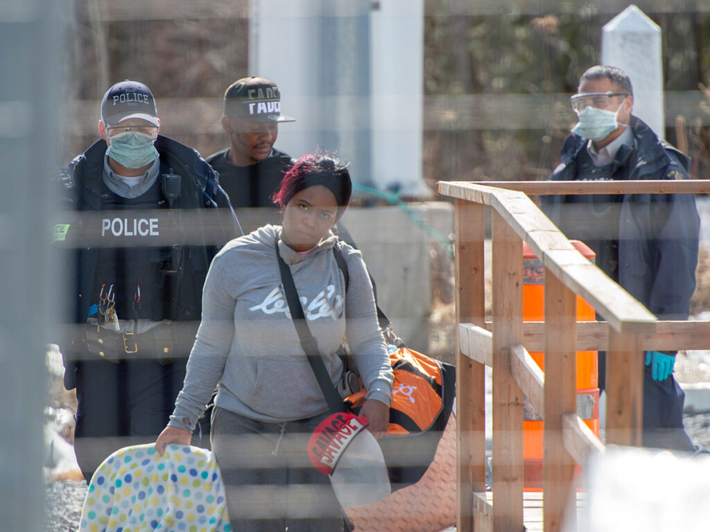 A woman holding a baby carrier and a bag over her shoulder is photographed with police in the background