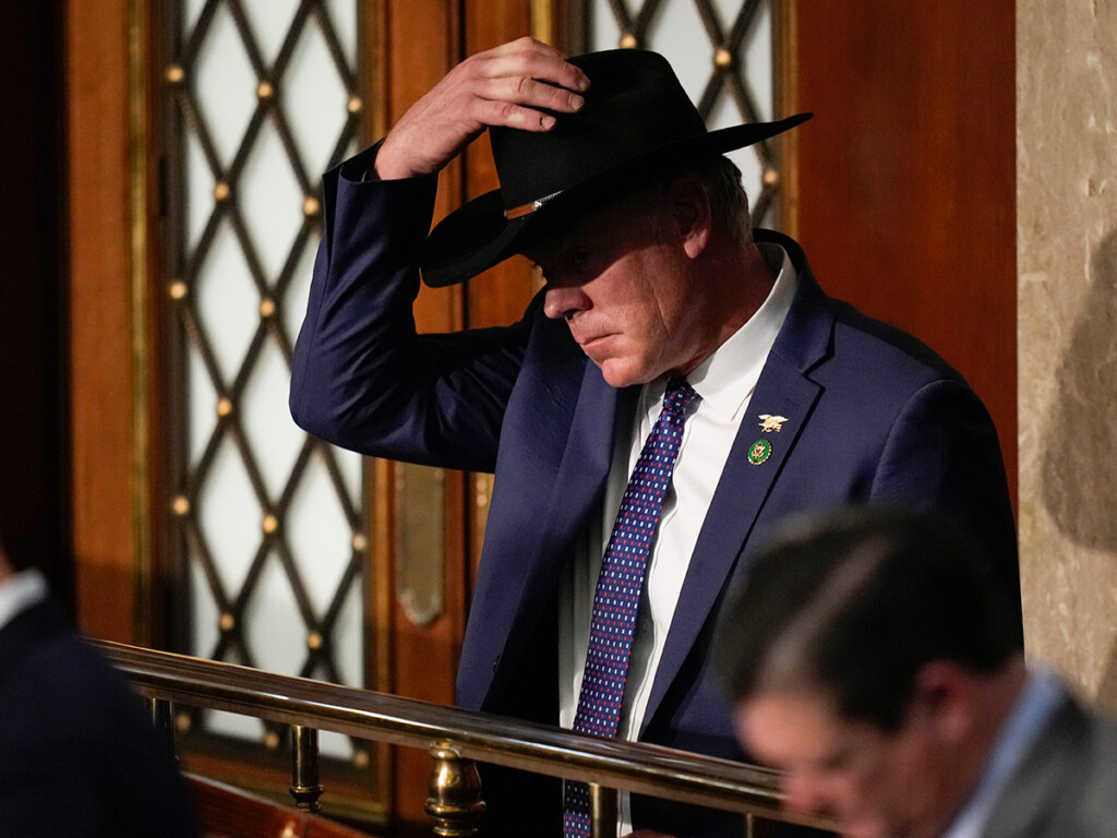 A man in a business jacket holds a cowboy hat on his head.