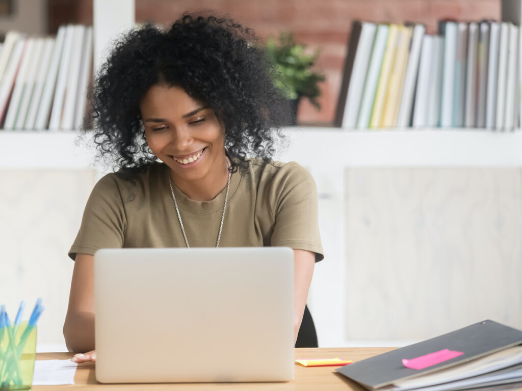 A woman sitting at a desk smiles at her laptop.