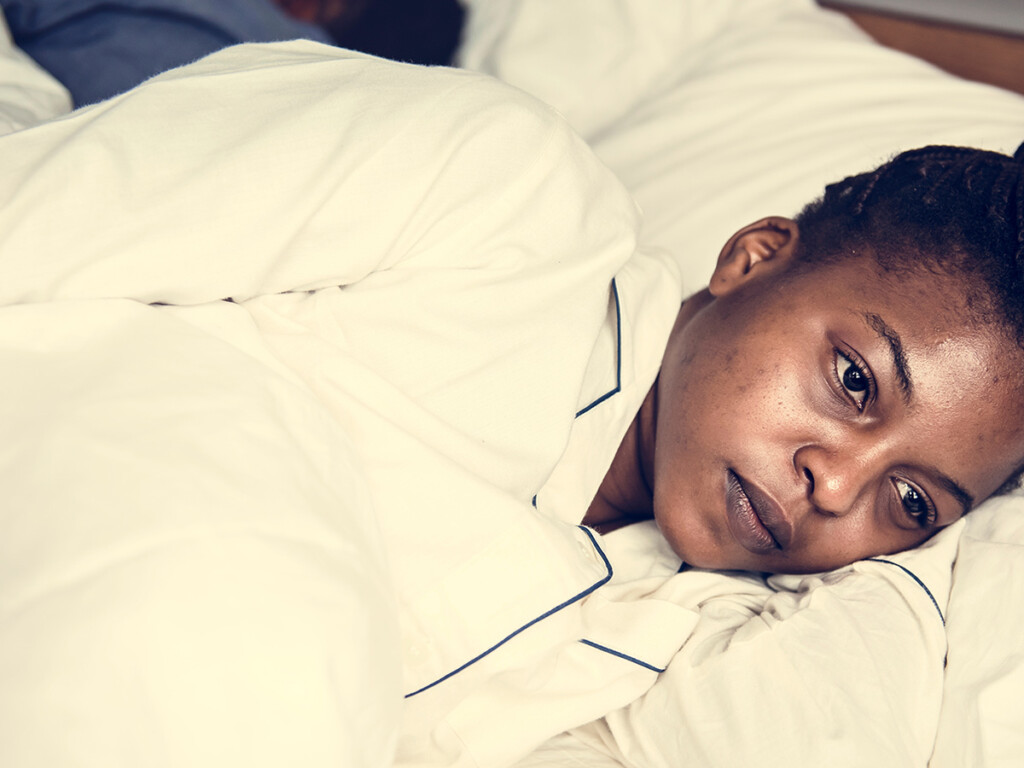 A woman lays in bed on her side, while another lays on the bed next to her facing the other way