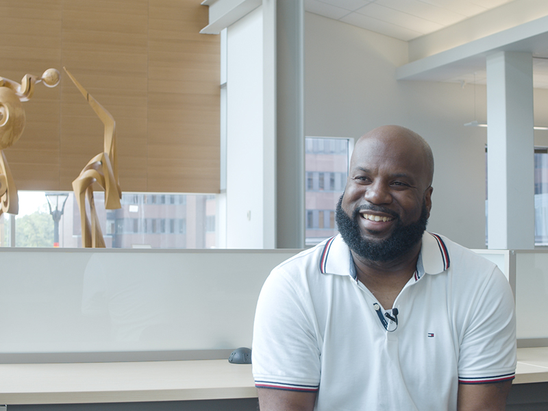 A man with a beard sits in front of an art gallery display, smiling for the camera