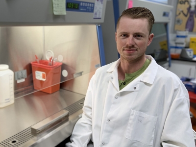 David Roy stands in a lab setting surrounded by equipment while wearing a lab coat.