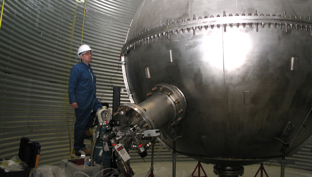 A man in a blue safety suit and white hard hat examines a large piece of equipment used for researching dark matter.