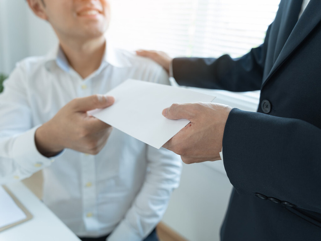 In an office setting, one man hands another man a white envelope.