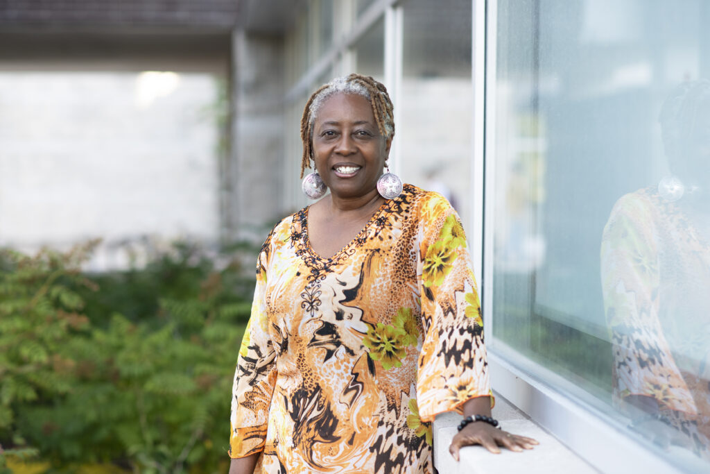 A woman woman stands outside and poses for a photo while resting her hand on a window sill.