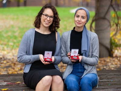 Mercedes Veselka and Samah Saci hold their medals outdoors on a bench in Alumni Park with fallen leaves in the background.