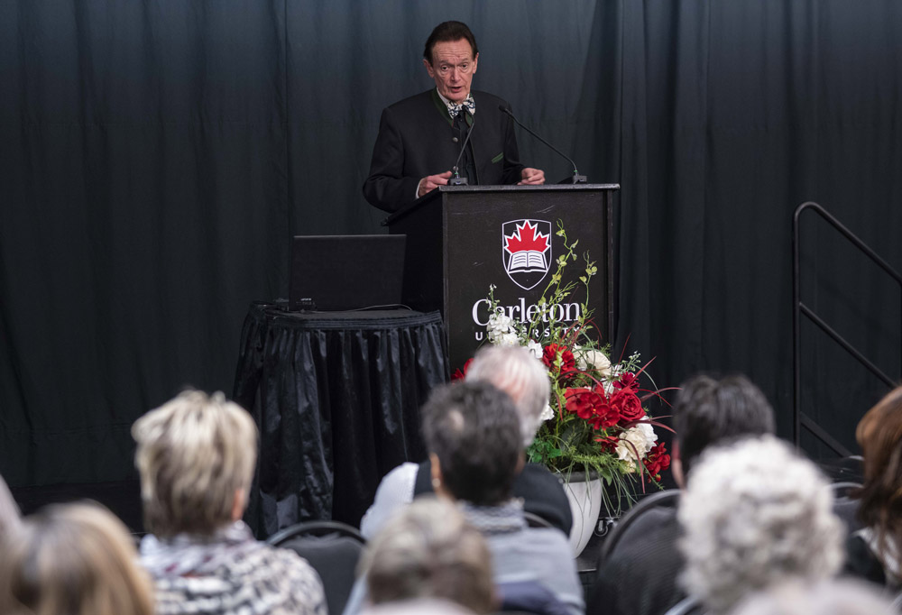 Prof. Martin Kemp speaks at a podium about the Salvator Mundi during the Herzberg Lecture while an audience listens.