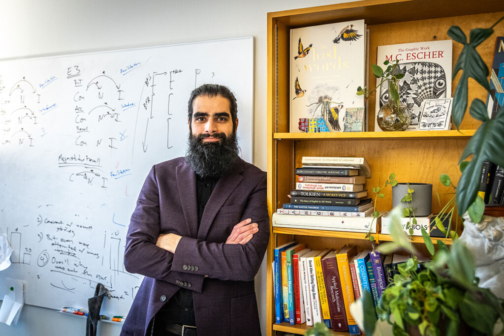 A man with a dark beard, wearing a purple dress coat, poses with his arms crossed next to a bookshelf.