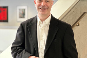 A man with a black dress suit and white buitton up shirt rests his arm on a banister while smiling for the camera.