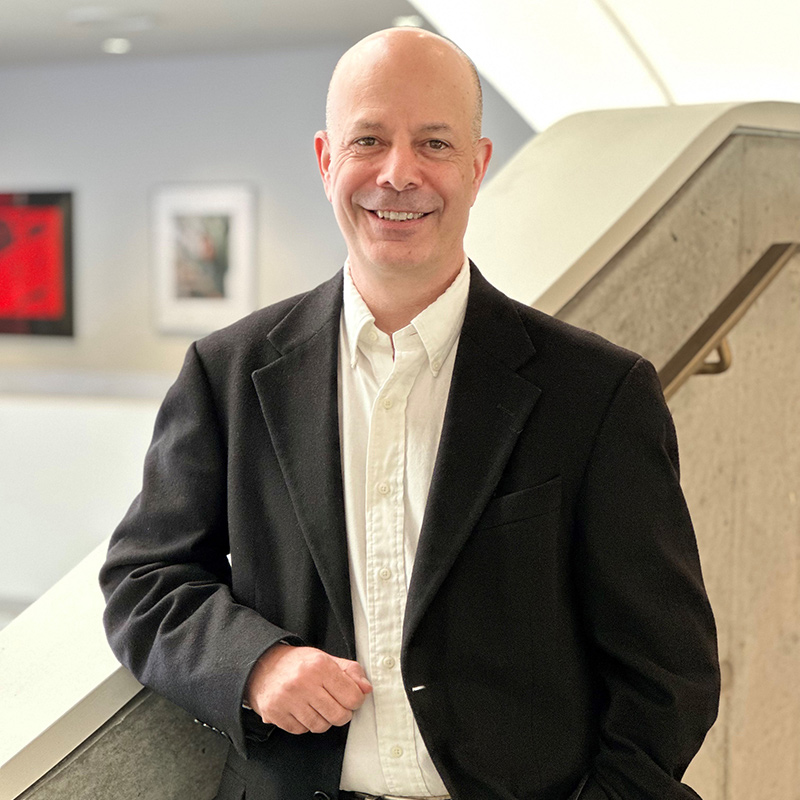 A man with a black dress suit and white buitton up shirt rests his arm on a banister while smiling for the camera.