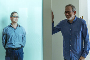 A man rests his hand on a holopresence unit project the full body image of another man.