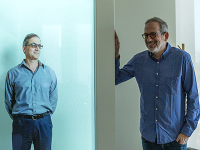 A man rests his hand on a holopresence unit project the full body image of another man.