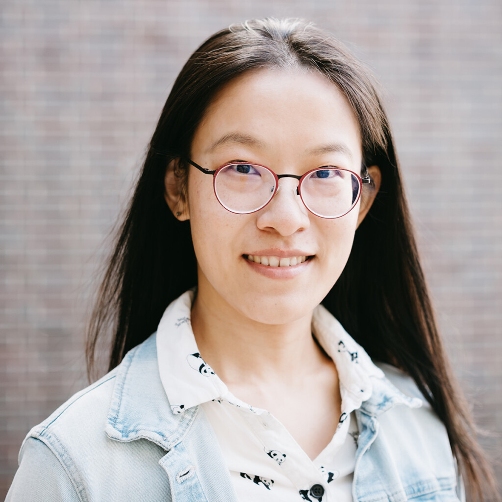 A young woman with glasses and wearing a jean jacket smiles for the camera