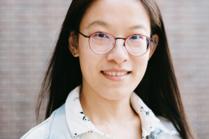 A young woman with glasses and wearing a jean jacket smiles for the camera