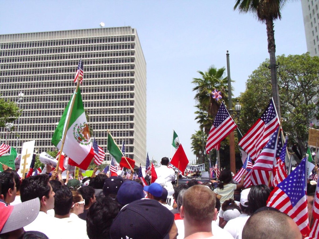 Large crowd at an immigration protest holding Mexican and American flags in a city street with tall buildings and palm trees in the background.