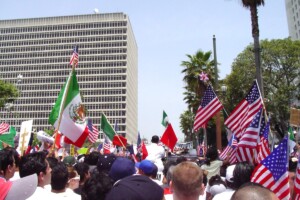 Large crowd at an immigration protest holding Mexican and American flags in a city street with tall buildings and palm trees in the background.