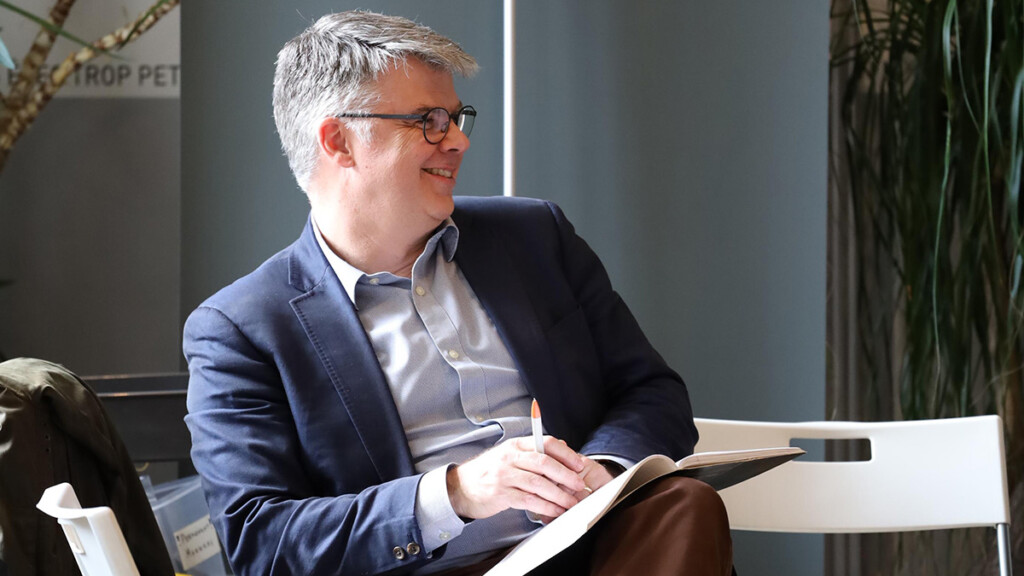 A professionally dressed man wearing glasses looks away from the camera while holding a pen and a book.