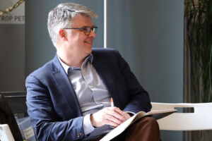 A professionally dressed man wearing glasses looks away from the camera while holding a pen and a book.