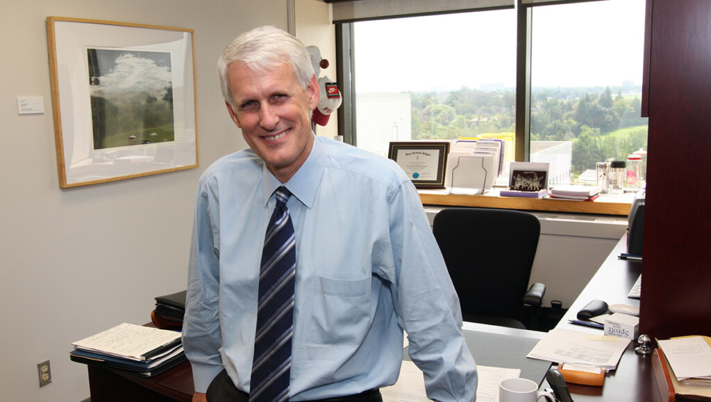 A man wearing a dress shirt and tie leans against a desk while posing for a photo.
