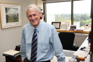 A man wearing a dress shirt and tie leans against a desk while posing for a photo.
