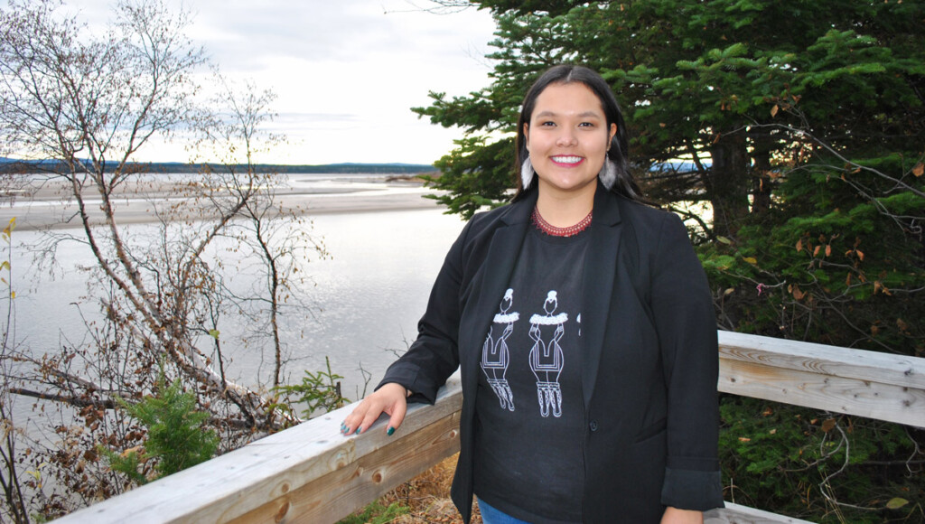 Jessica Lyall stands outdoors on a wooden deck situated next to a northern Canadian lake with an evergreen tree behind her.
