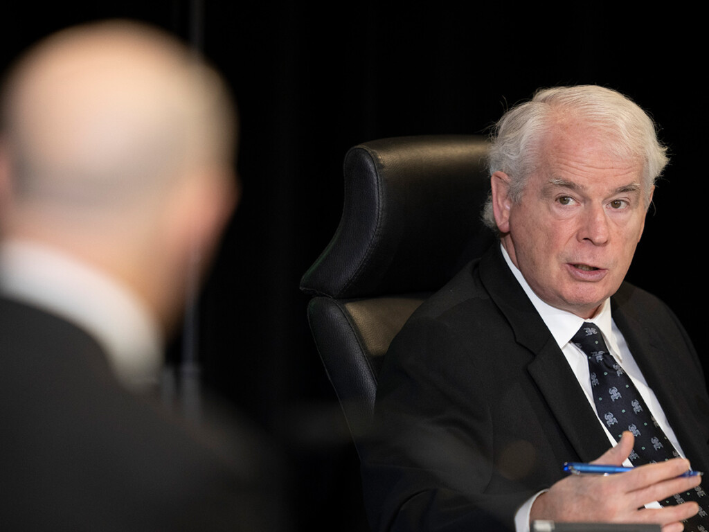 A man with grey hair sits behind a desk speaking to someone seen from behind in the foreground.