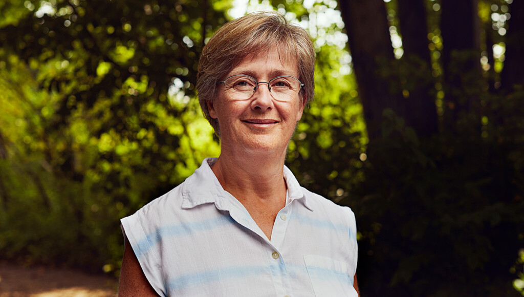 A woman with short hair wearing glasses smiles at the camera with trees behind her.