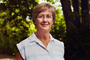 A woman with short hair wearing glasses smiles at the camera with trees behind her.