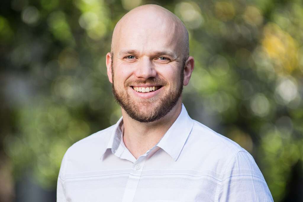 A man who is bald with a beard, wearing a white collared shirt smiles for the camera