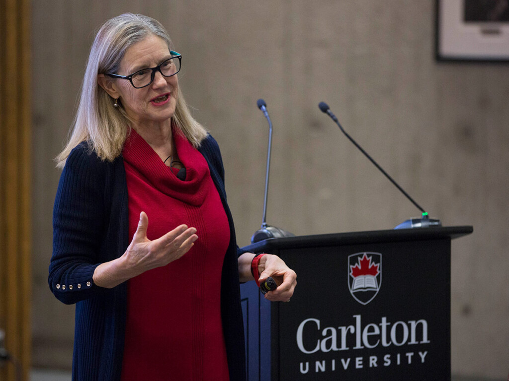A woman with glasses wearing a red shirt and blue cardigan stands next to a podium with a microphone to deliver a speech.