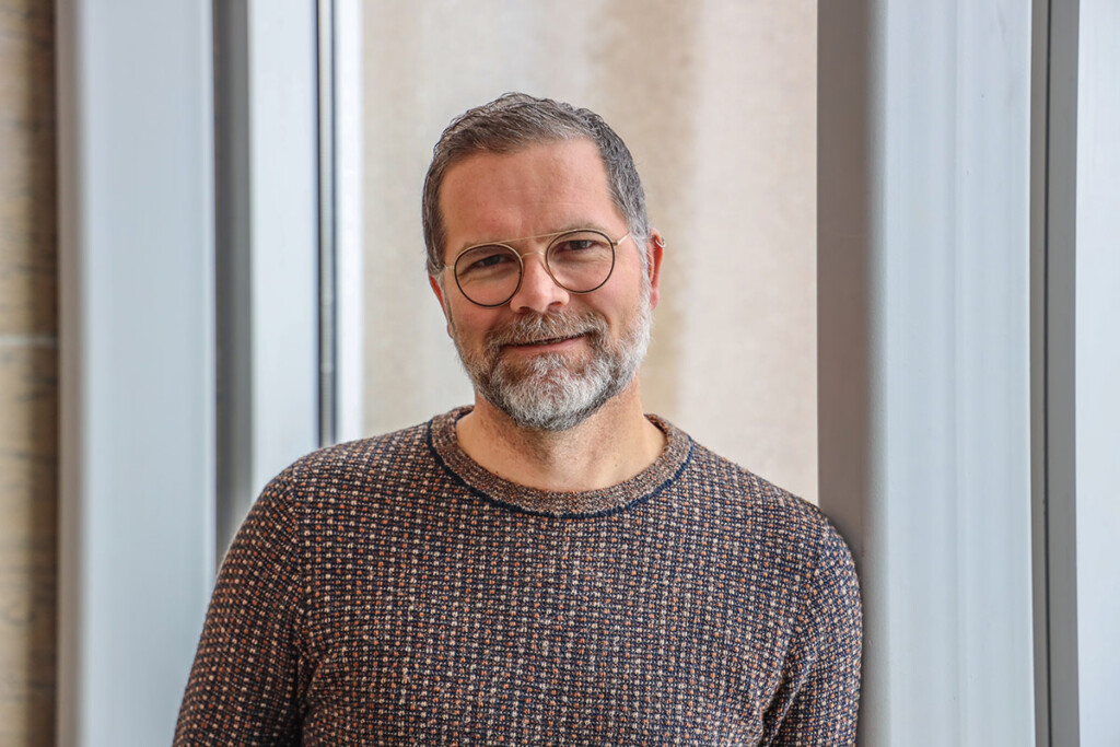 A man wearing glasses smiles for the camera while standing in front of a window.