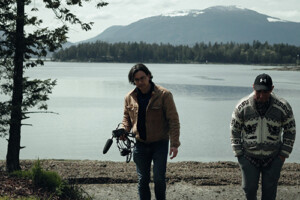 Two men, including one holding a camera, walk away from the shore of a lake.