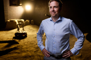 A man in a button-up shirt looks away from the camera, a lunar rover is seen in the background.
