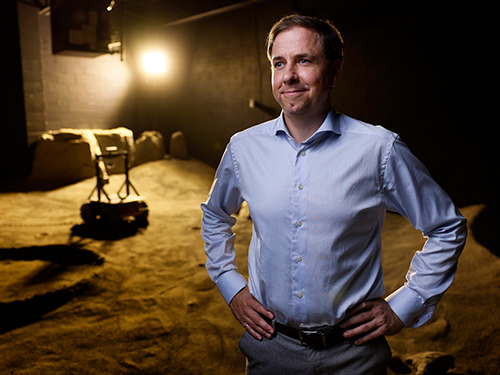 A man in a button-up shirt looks away from the camera, a lunar rover is seen in the background.