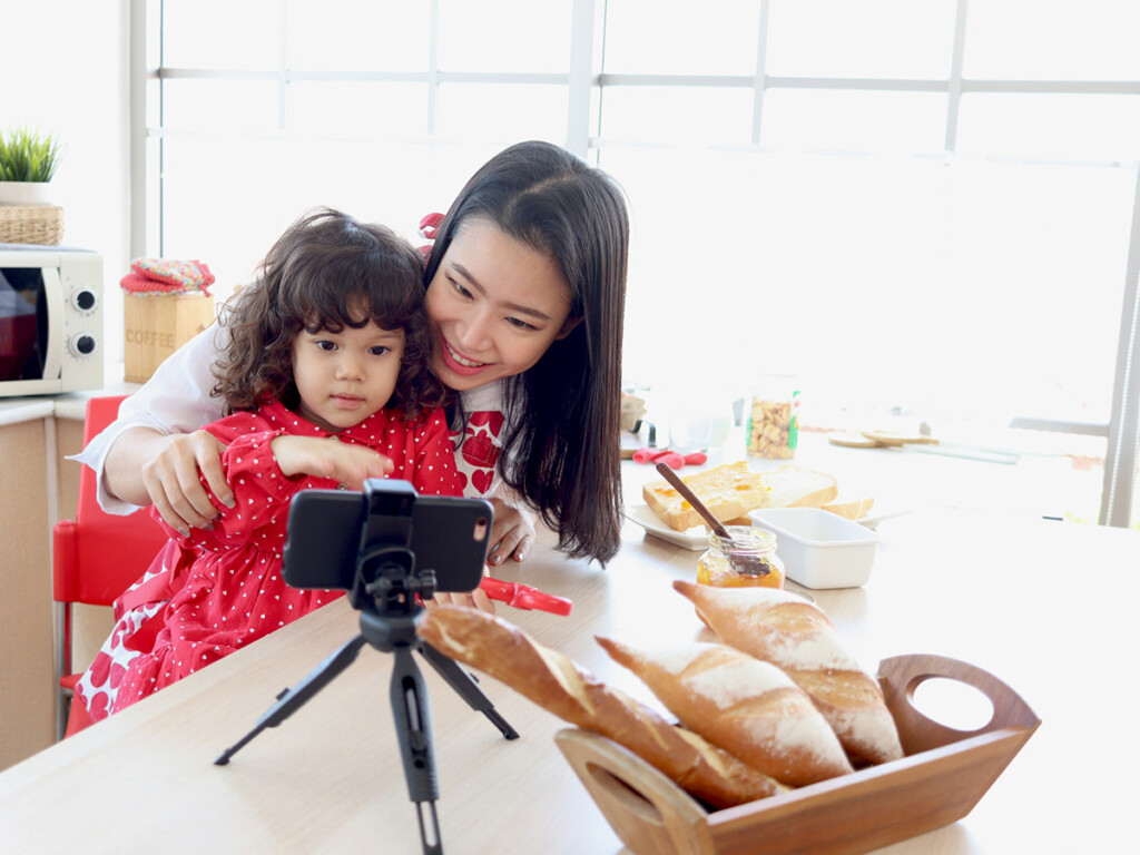 A mother and child recording a video on a mobile phone in their kitchen. Image is intended to depict momfluencers on social media.