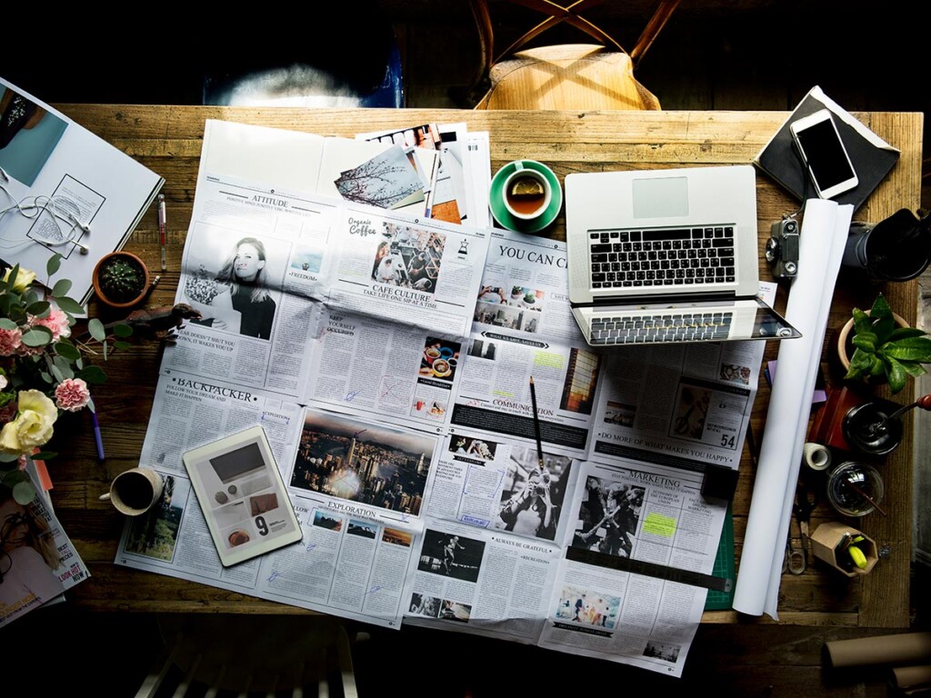 A newspaper spread out on a wooden table with a cup of coffee and a laptop resting on top.