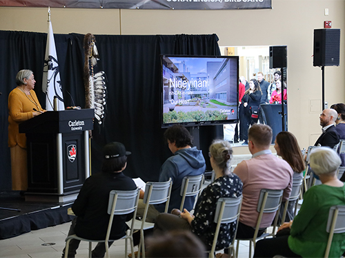 A woman addresses a crowd from a podium across from a large screen