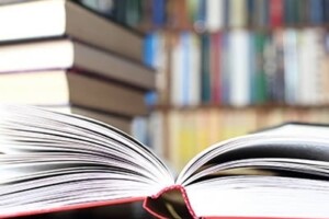 An open book on a wooden table. A stack of books sits behind it.