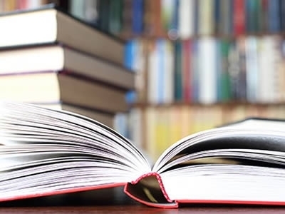 An open book on a wooden table. A stack of books sits behind it.