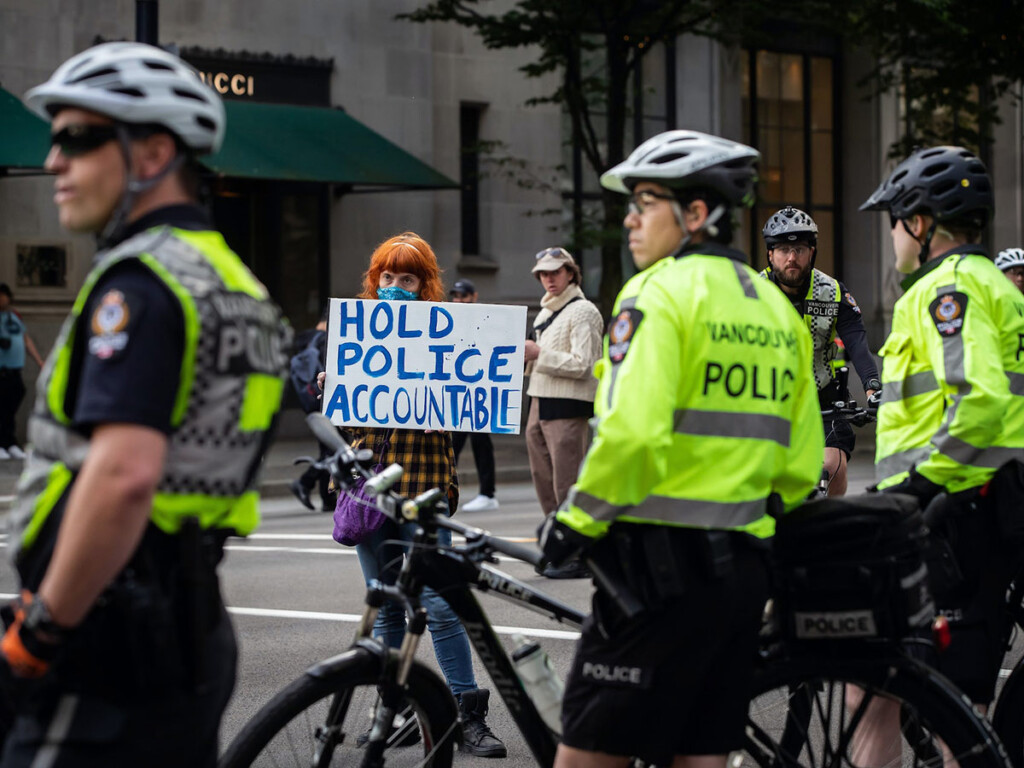 Four Vancouver Police officers stand on a street with a protestor visible in the distance holding a sign that reads: Hold Police Accountable