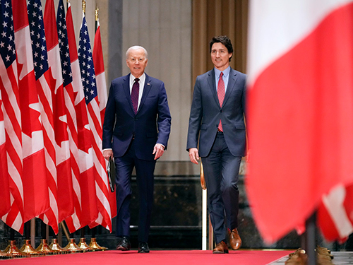 President Biden and Prime Minister Trudeau walk together along a red carper lined with American and Canadian flags.