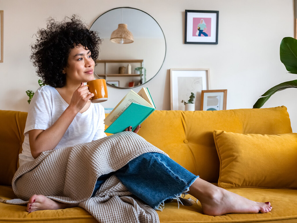 Pensive relaxed woman reading a back-to-school stress help book at home, drinking coffee sitting on the couch.