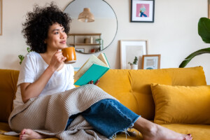 Pensive relaxed woman reading a back-to-school stress help book at home, drinking coffee sitting on the couch.