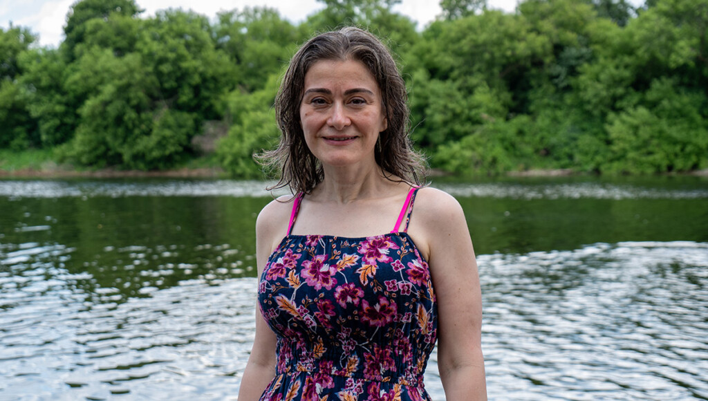 A woman wearing a floral bathing suit poses for the camera in front of a river and trees.