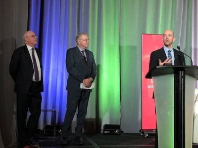 Carleton President Benoit-Antoine Bacon speaks at the podium during the Royal Society Celebration while two men in suits watch on.