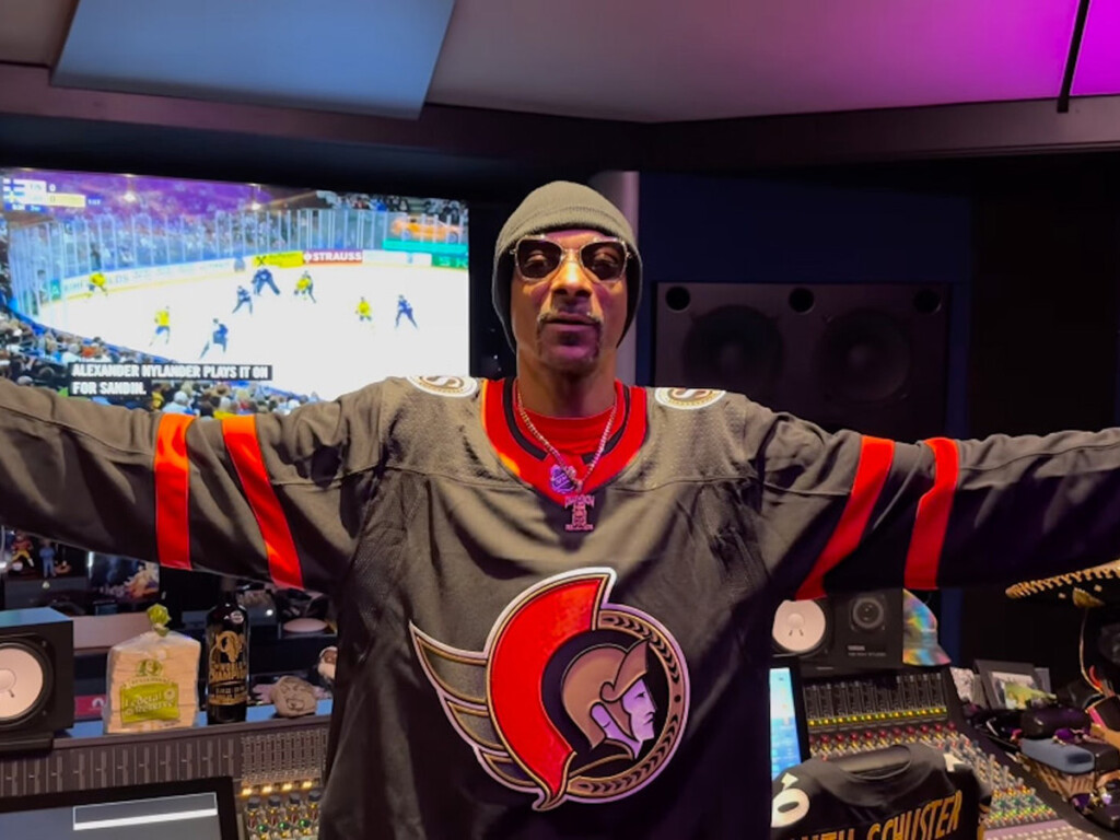 A man in a black toque and hockey jersey stands with open arms in front of a TV displaying a hockey game.