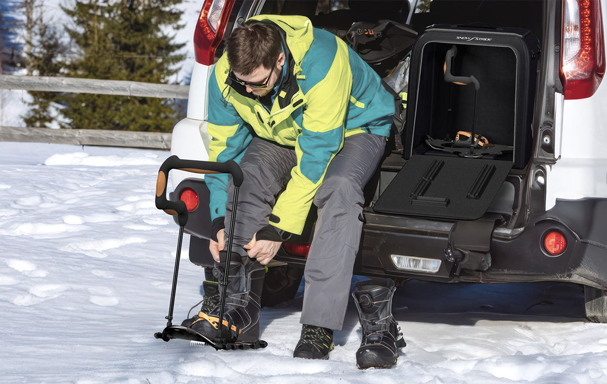 A man sits on the back of his car while putting on a SnowStride snowshoe-inspired mobility aid.
